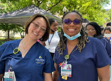 Nurses Mayumi Shigeno and Lajuana Willis, wearing blue scrubs and smiling, with the tents of the farmers’ market behind them. 